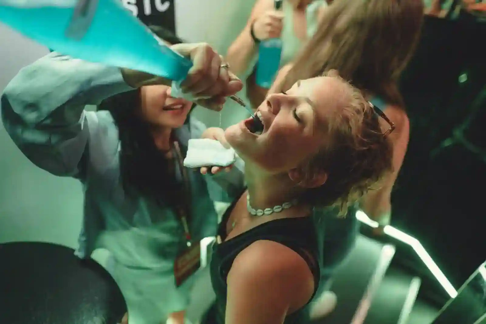 yc8ustcnde5yv2eusy3f_compressed_compressed A woman tilts her head back as another person pours a blue liquid into her mouth at what appears to be a social gathering.