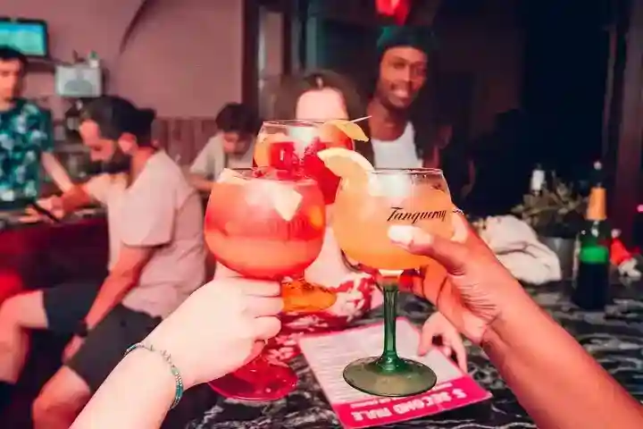 khiybjyfrsplomnthxoe_compressed_compressed Three people clink colorful cocktails together in a lively bar, with other patrons and drinks visible in the background.