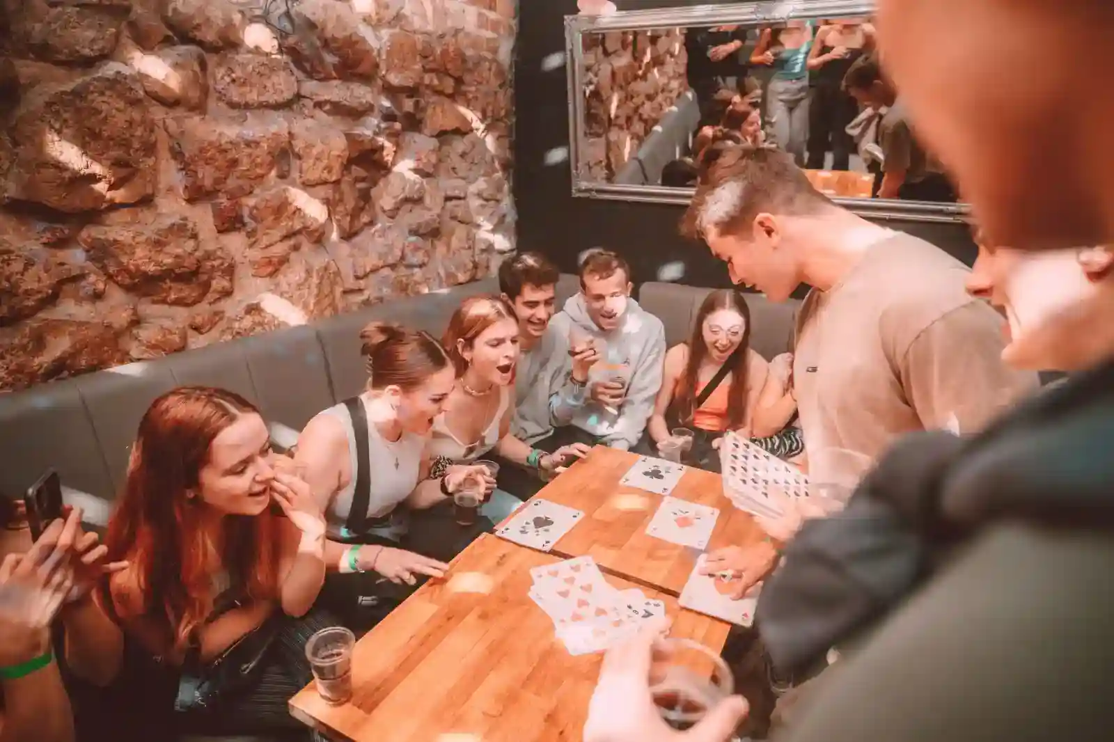 xzchknqpkekxh5rbedmd_compressed_compressed A group of people sitting around a table react excitedly as a man performs a card trick in a cozy, stone-walled room.