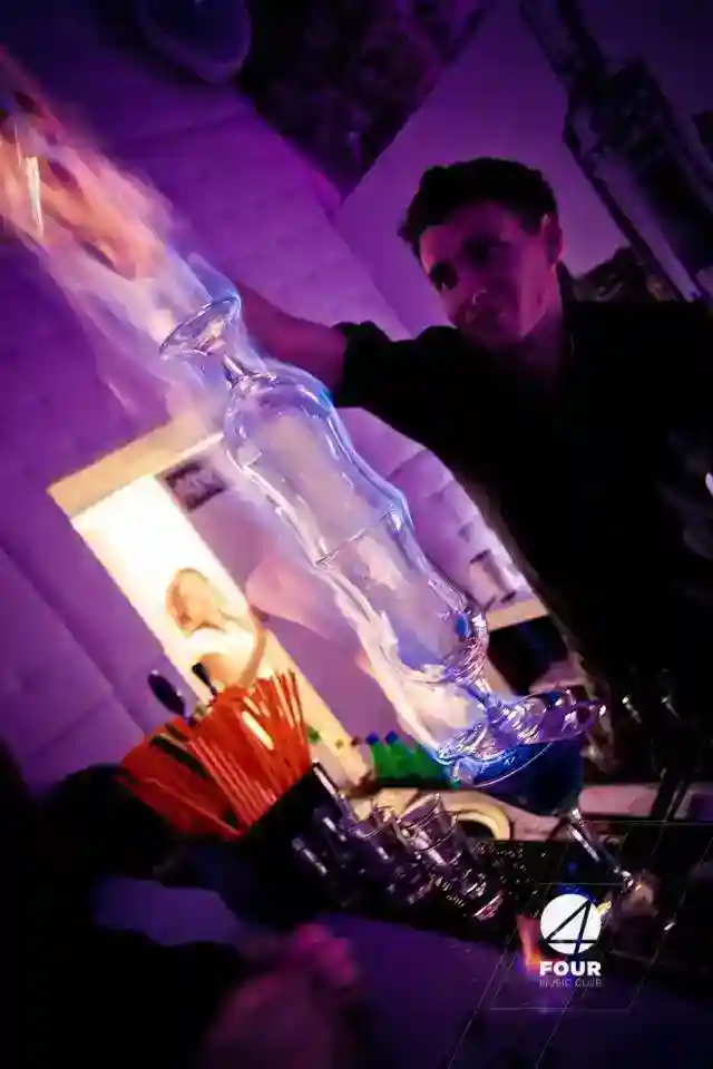 A bartender pours blue liquid through a tall, stacked glass tower at a bar with purple lighting and cocktail straws visible in the foreground.