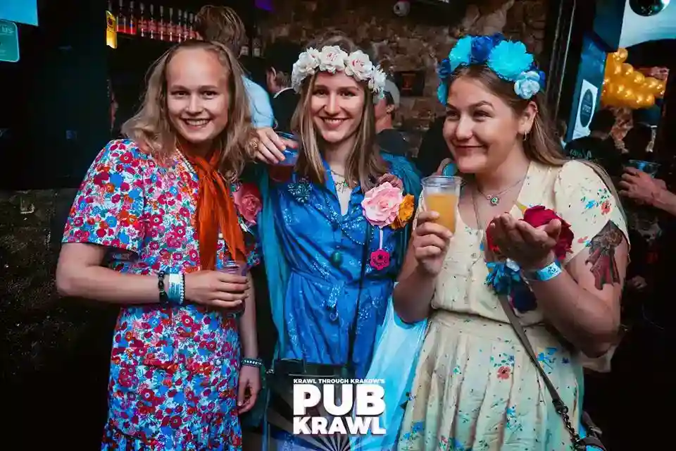 Three women wearing floral dresses and flower crowns hold drinks and smile at a pub crawl event. "Pub Krawl" logo is visible at the bottom of the image.