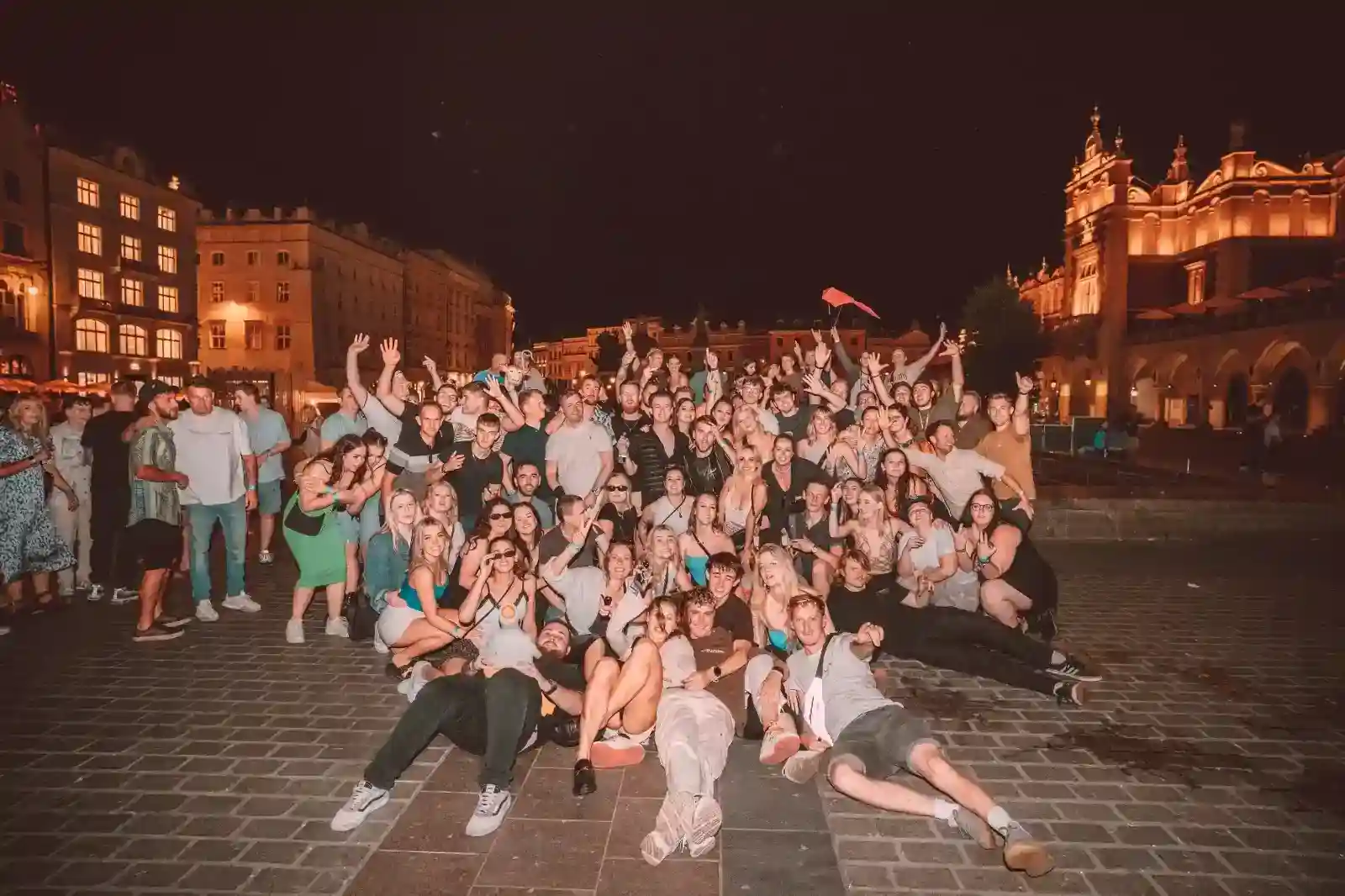 qjfkytgsbnnjmkmkcfrc_compressed_compressed A large group of people pose together at night in a city square with historic buildings and lights in the background.