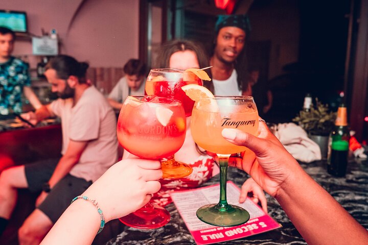 Close-up of three people clinking their colorful cocktail glasses at a bar during a Krakow pub crawl. Other patrons and items are visible in the background.